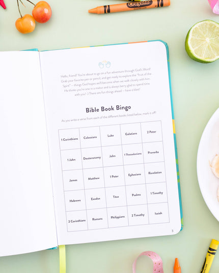 Open book with 'Bible Book Bingo' game on a table with snacks and stationery items.