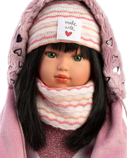 Doll wearing a pink hooded coat, striped hat, and scarf with a heart label.