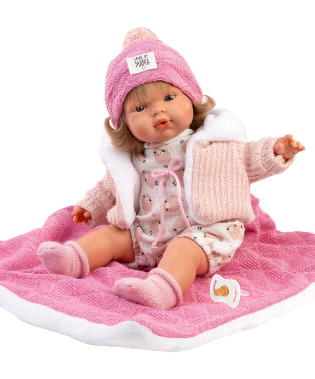 Baby doll in pink outfit with matching hat on a pink blanket against a white background