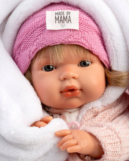 Baby doll wearing a pink knitted hat with 'MADE BY MAMA' label, wrapped in white blanket.