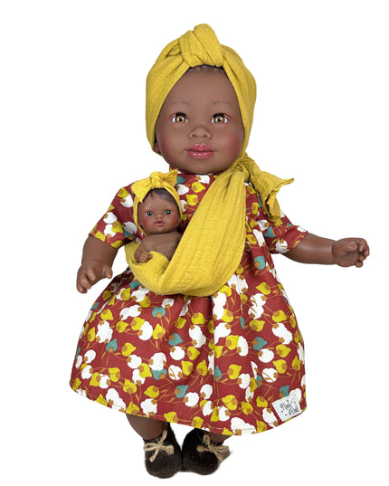 Doll wearing a yellow headscarf and floral dress holding a smaller doll wrapped in a yellow blanket on a white background.