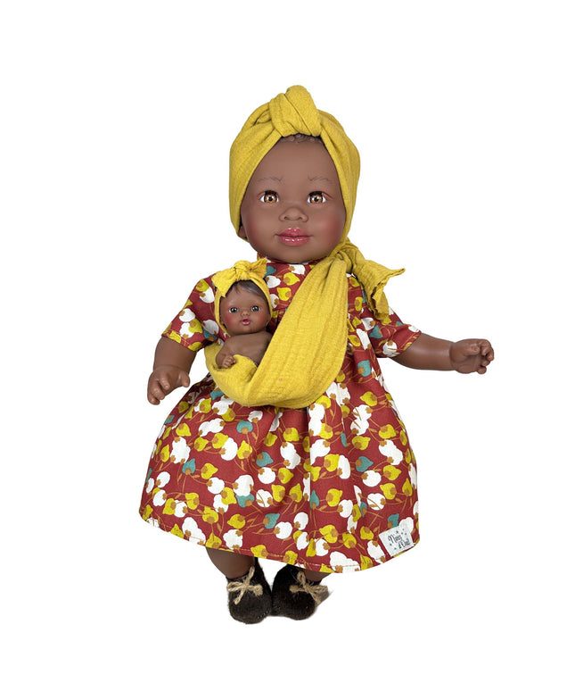 Doll wearing a yellow headscarf and floral dress holding a smaller doll wrapped in a yellow blanket on a white background.
