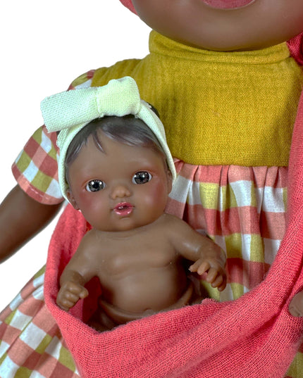 Doll holding a baby doll wrapped in a pink blanket with a checkered dress in the background