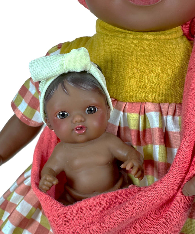 Doll holding a baby doll wrapped in a pink blanket with a checkered dress in the background