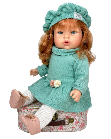 Doll in a teal outfit with a hat sitting on a floral suitcase against a white background