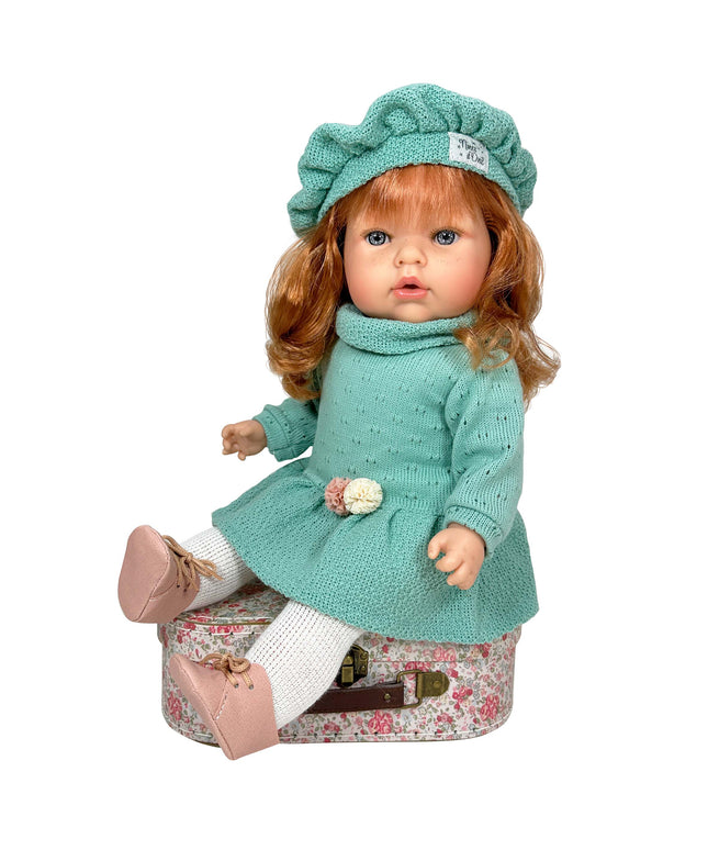 Doll in a teal outfit with a hat sitting on a floral suitcase against a white background