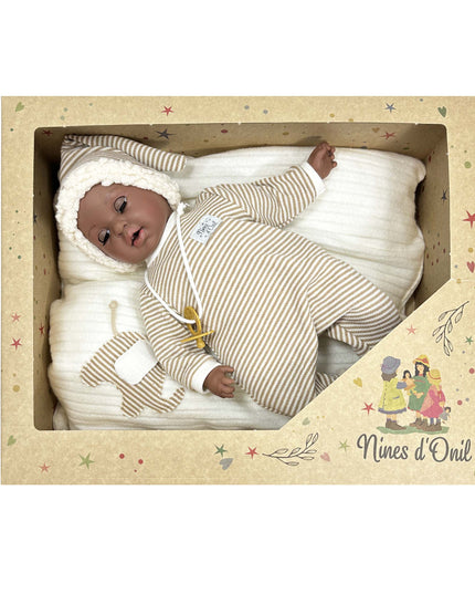 Baby doll in a striped outfit and white hat lying on white fabric inside a decorative box with 'Nines d'Oril' branding.