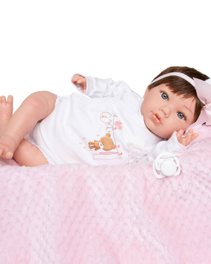 Baby doll with brown hair and blue eyes wearing a white outfit with a teddy bear design, lying on a pink textured surface.
