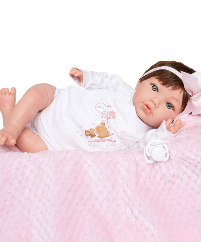 Baby doll with brown hair and blue eyes wearing a white outfit with a teddy bear design, lying on a pink textured surface.