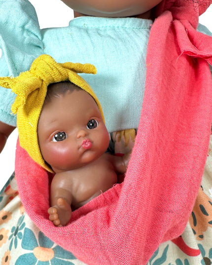Baby doll with a yellow bow in a pink sling held by a person wearing a floral dress.