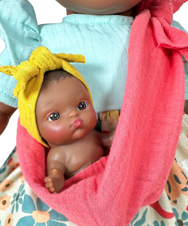 Baby doll with a yellow bow in a pink sling held by a person wearing a floral dress.