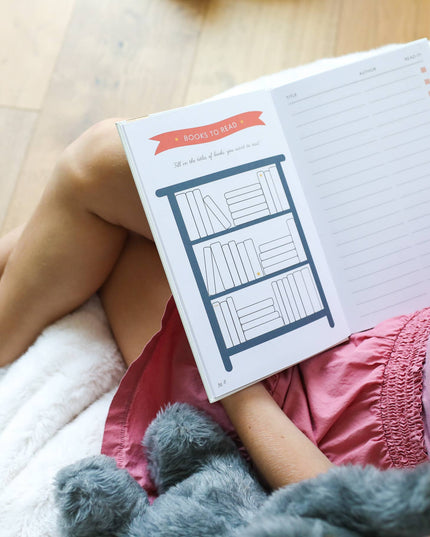 Person lying on a wooden floor with a book covering their face, wearing pink pants and a gray sweater.