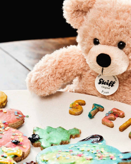 Teddy bear with a Steiff tag next to colorful cookies shaped like letters on a white plate.