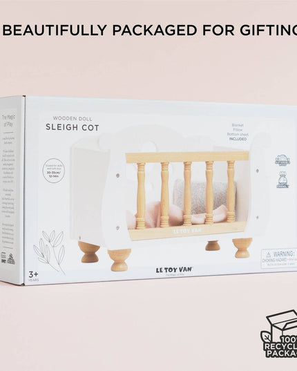 Packaged wooden doll sleigh cot with branding and recyclable packaging label on a light pink background