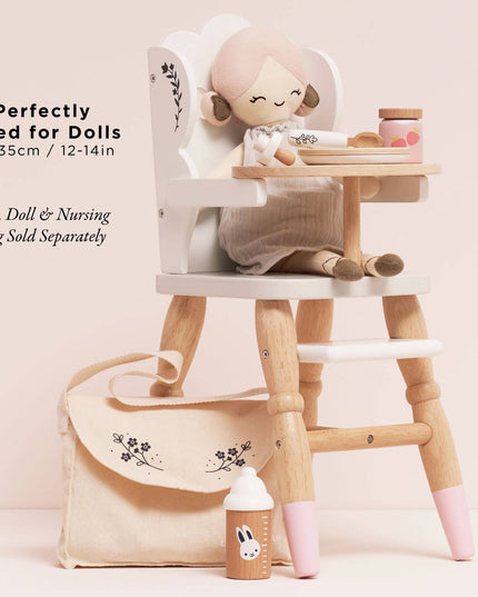 Wooden doll high chair with a doll on top, accompanied by a bag and bottle on a light pink background.