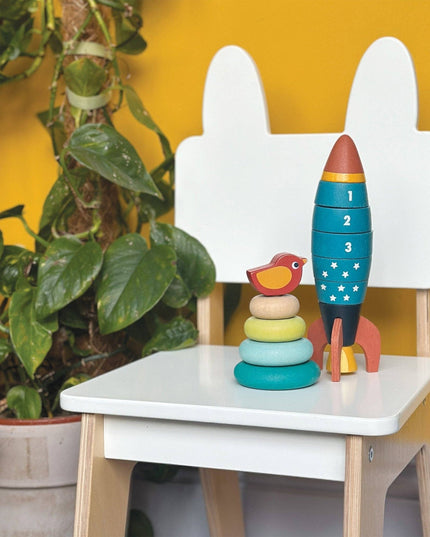 Colorful toy rocket and wooden stacking rings on a white table with a yellow wall and plant in the background