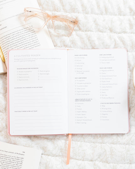 Open book with a pink cover on a white textured surface, with a cup of tea and glasses in the background.
